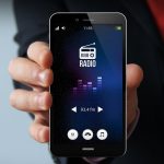 A close-up photo of a man's hand, holding a smartphone, with the screen displaying a radio broadcast app.