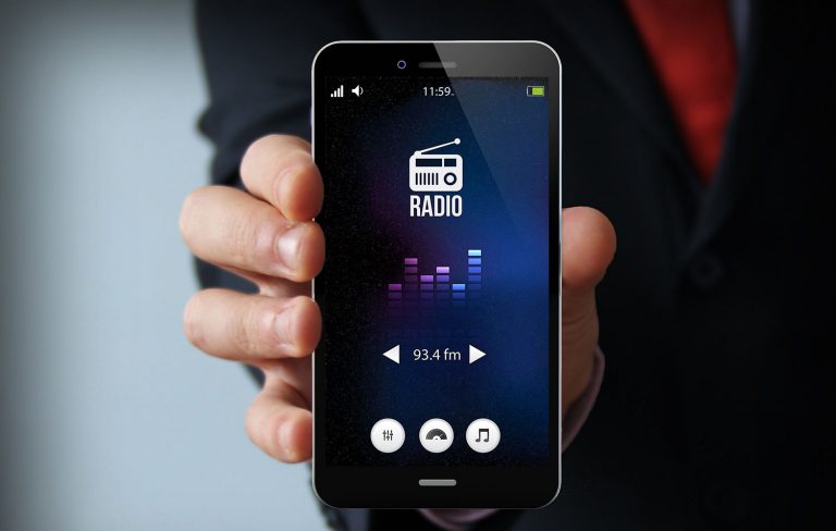 A close-up photo of a man's hand, holding a smartphone, with the screen displaying a radio broadcast app.